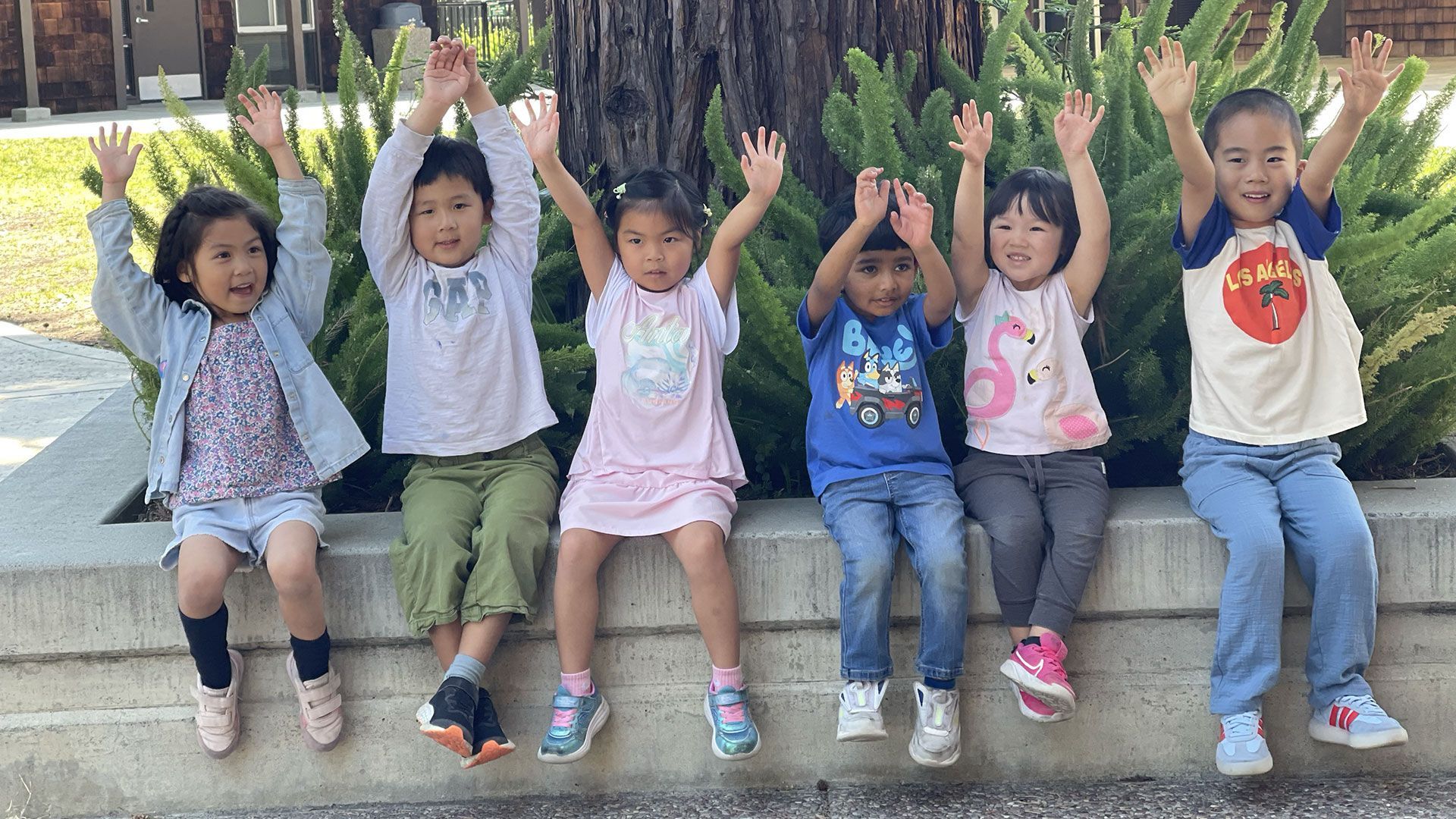a group of happy children sitting on a short cement wall with hands raised