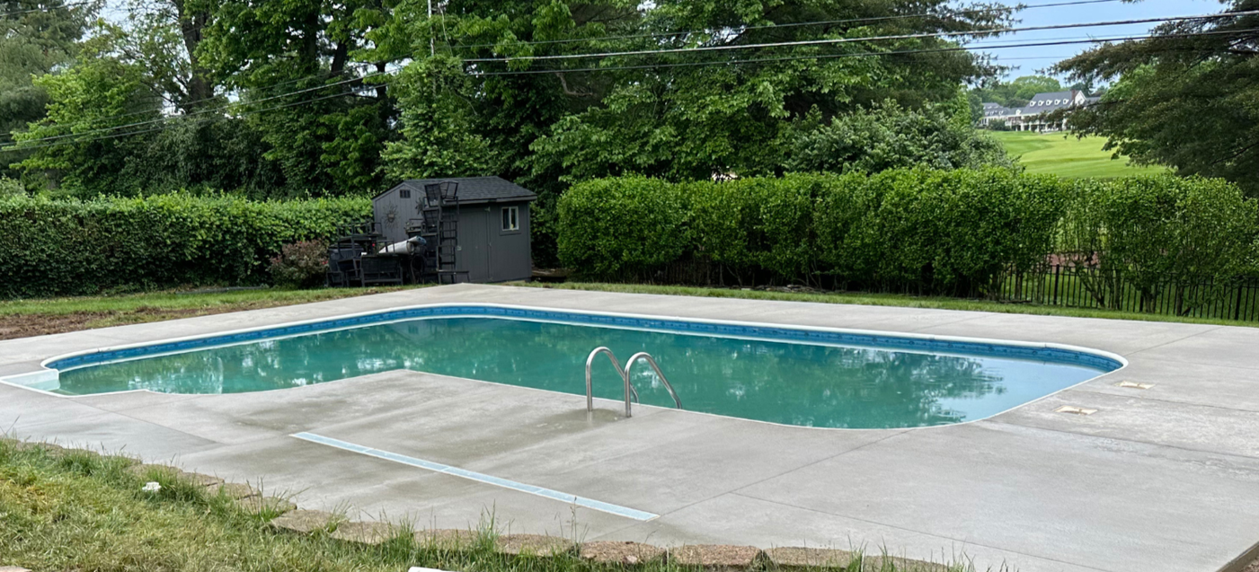 A rectangular swimming pool with blue trim, concrete deck, and green lawn, with a shed and trees in the background.