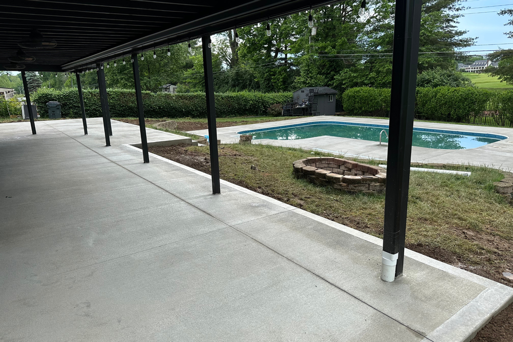 Covered patio next to a pool and fire pit. Concrete floor, black supports, green grass and trees.