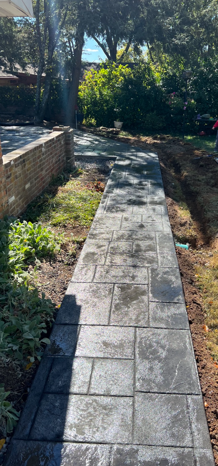 Stone pathway through a garden with a brick wall on the left and greenery in the background.