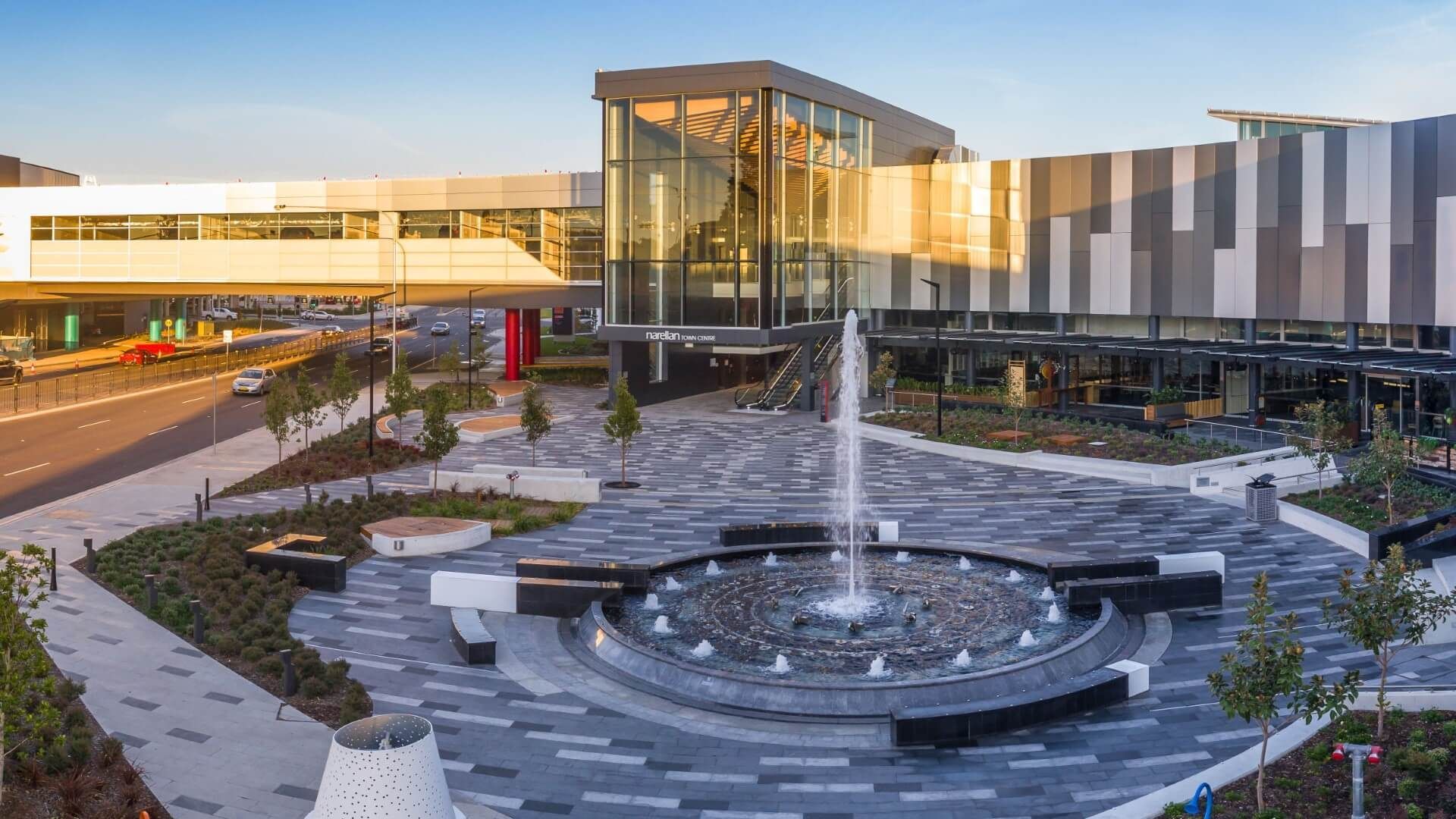 Exterior view of a modern shopping mall with a fountain in the foreground. The building has a mix of glass and gray/white paneling.