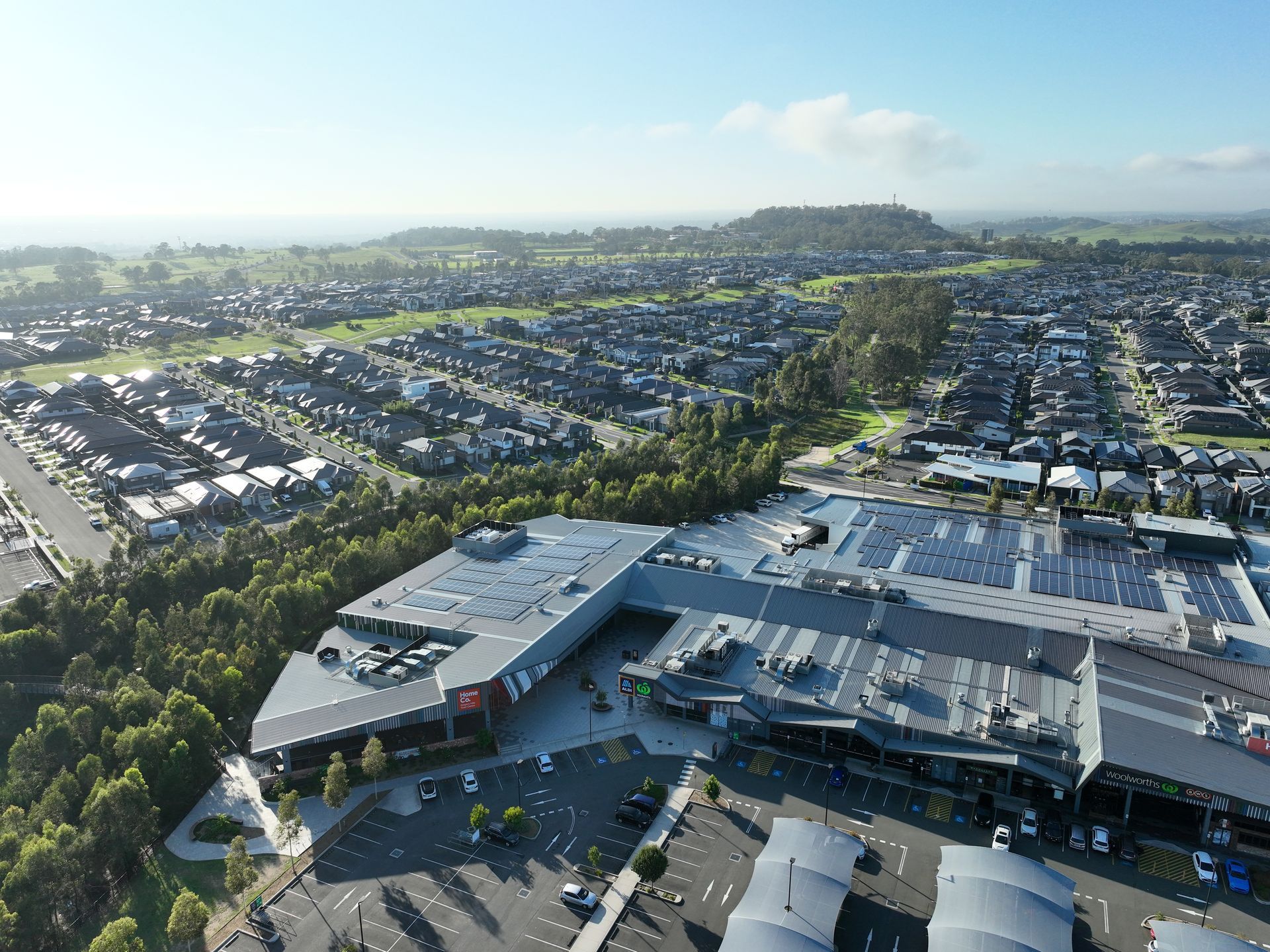 Aerial view of a shopping center with a large parking lot, surrounded by residential areas and green fields under a bright blue sky.