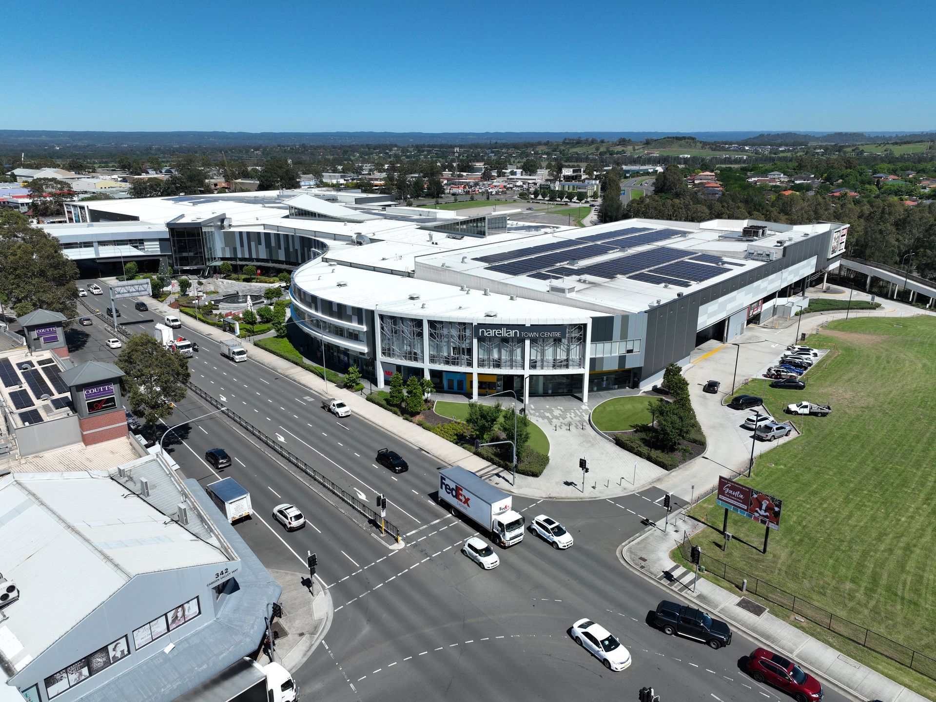 Aerial view of a large, modern building with solar panels on the roof, a curved glass facade, and a busy intersection in front.