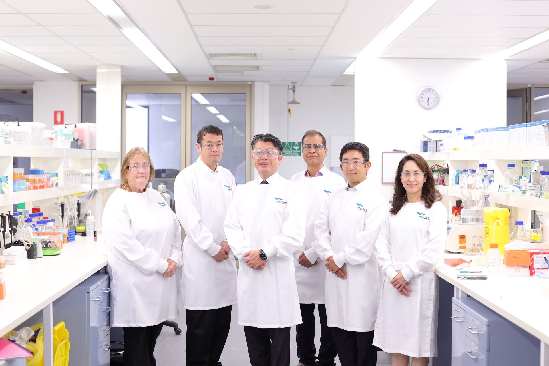 A group of six scientists in white lab coats stand in a laboratory setting, posing for a photo. Fluorescent lights and lab equipment are visible.