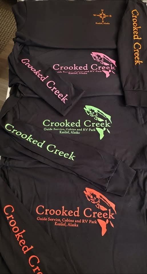 Stacked black T-shirts with “Crooked Creek” printed in pink, green, and orange on the front.