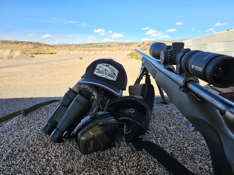 Binoculars and a rifle with scope on a shooting rest in a desert landscape.