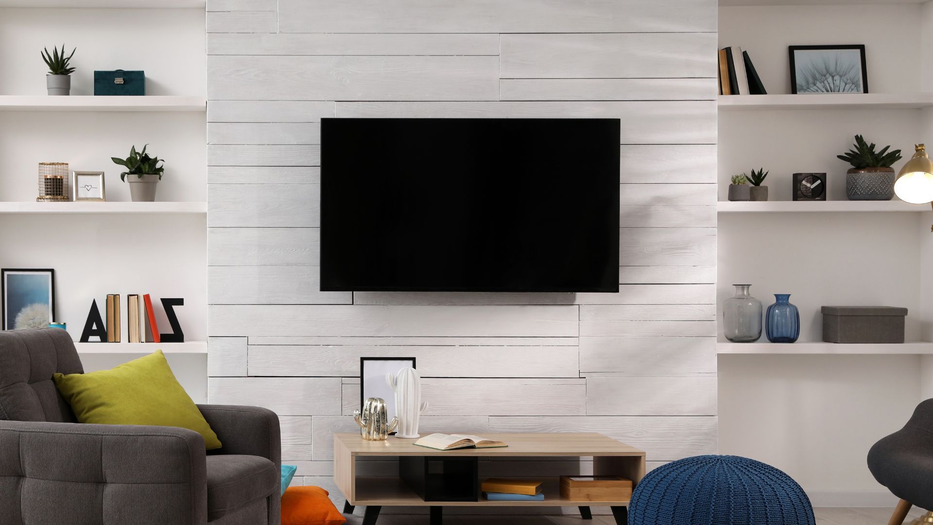 A modern living room with a wall-mounted television, wooden shelves filled with decor, and two armchairs.
