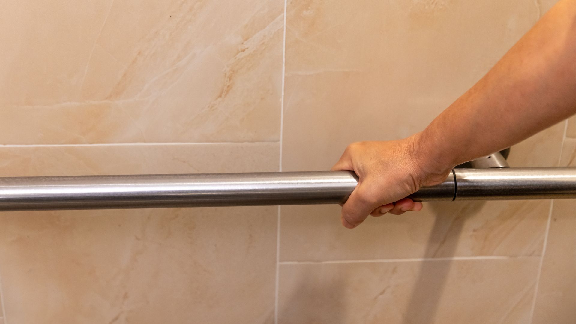 A hand gripping a horizontal stainless steel grab bar mounted on a beige tiled bathroom wall.