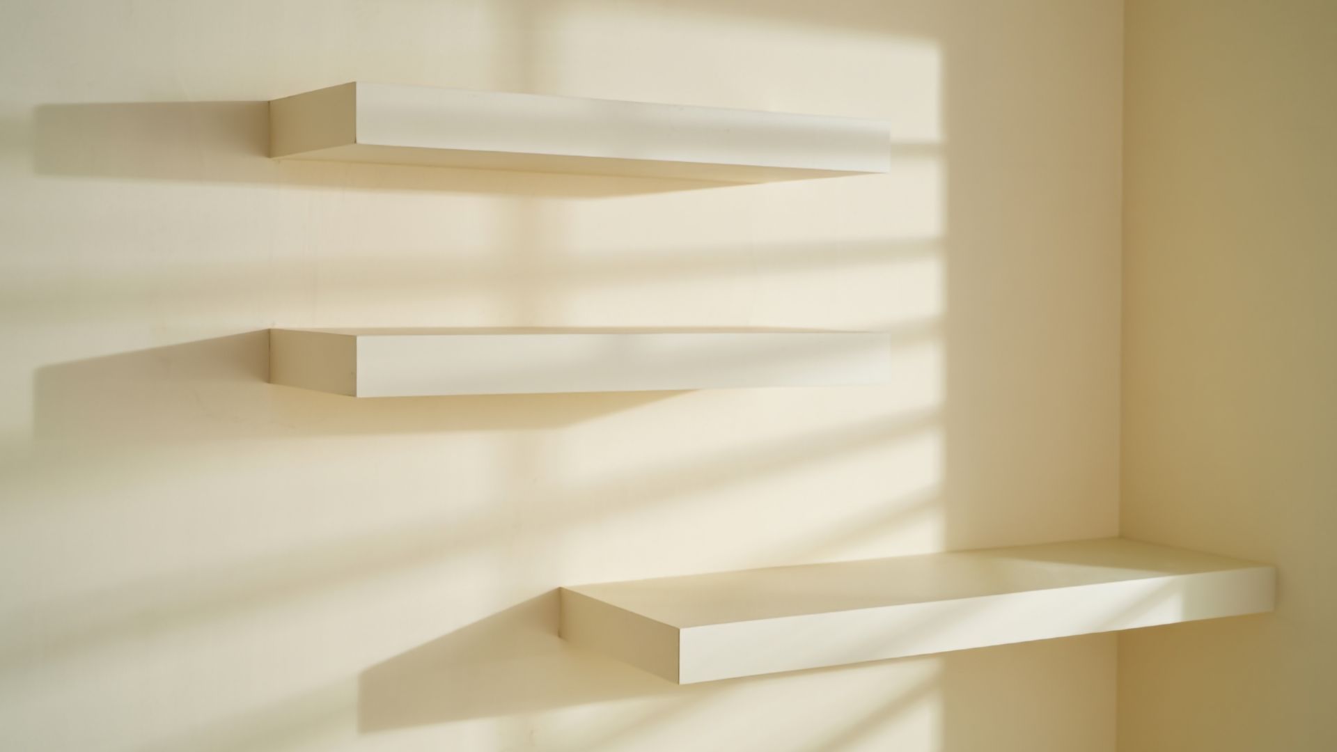 Three off-white floating shelves mounted against a plain wall with natural window light casting shadows.