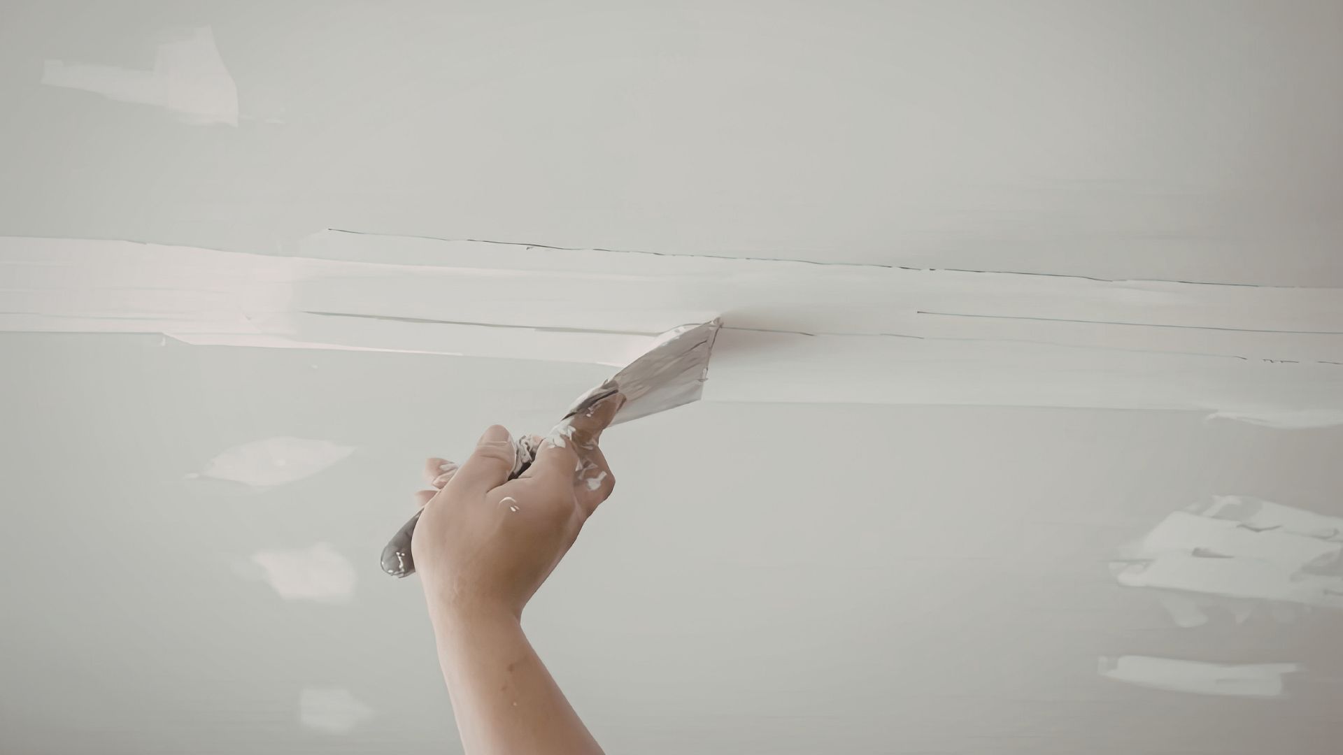A person uses a putty knife to spread white joint compound over a seam on a ceiling.