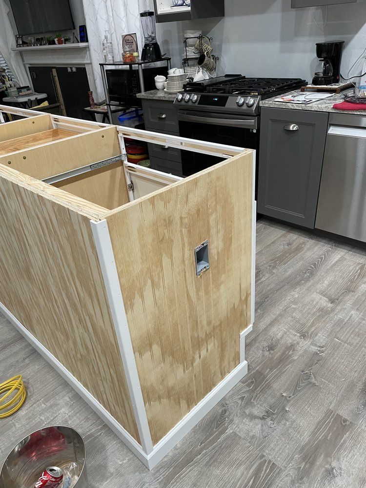 A plywood kitchen island frame with an electrical box cutout, installed on a wood-look floor near a kitchen stove.