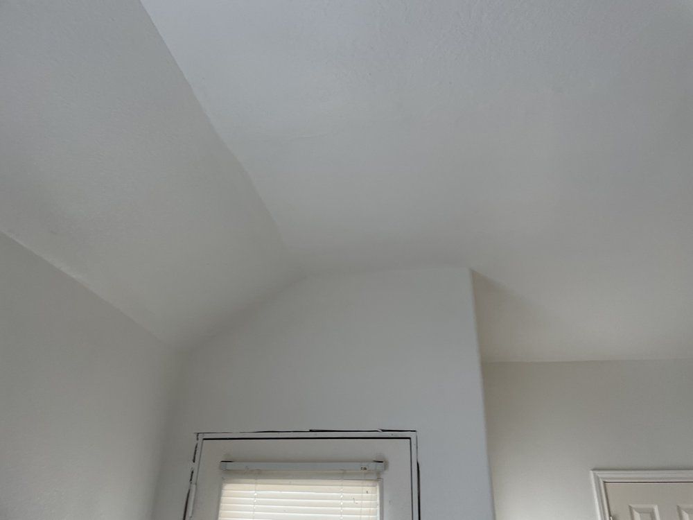 A low-angle view of a white, angled ceiling meeting a wall above a door frame with blinds.