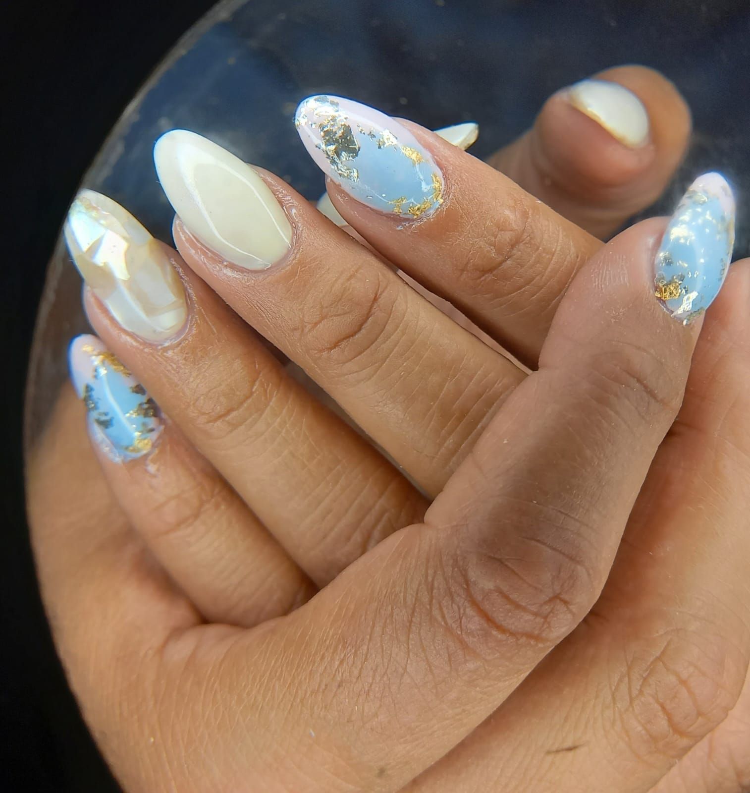 Manos con uñas almendradas de color azul claro y blanco, algunas con detalles en papel dorado.