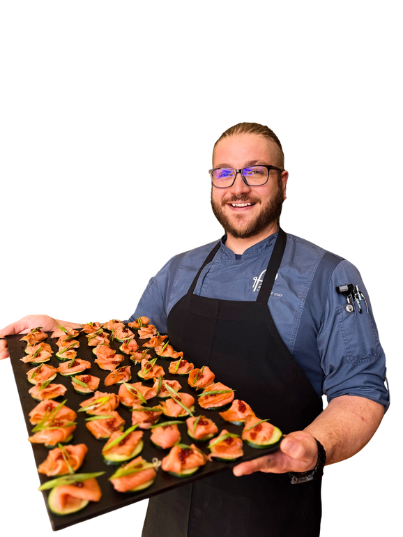 A smiling chef in a blue shirt and black apron holds a large rectangular tray of appetizers.