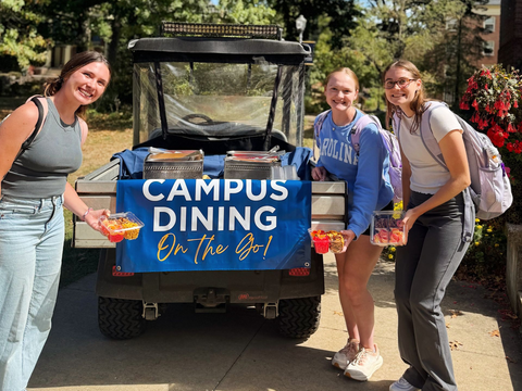 Three people stand smiling behind a Campus Dining golf cart, holding small containers of food.