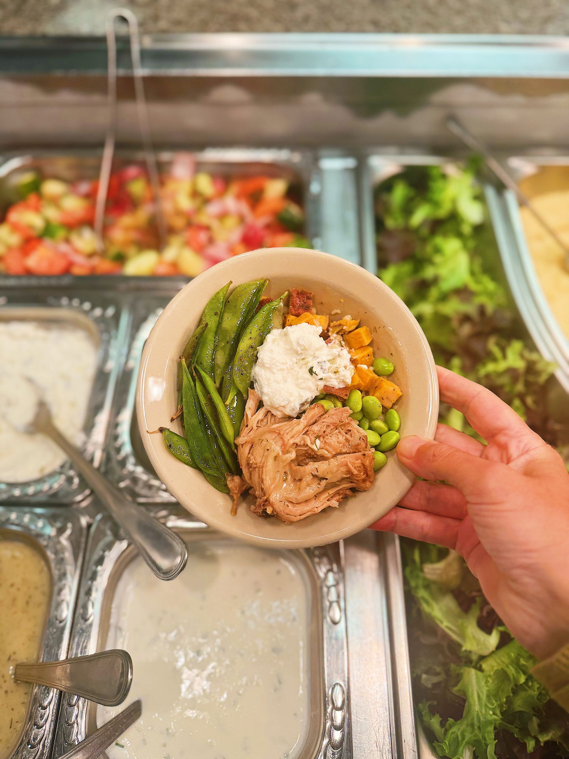 A hand holds a small bowl filled with shredded meat, snap peas, edamame, and a dollop of white sauce.