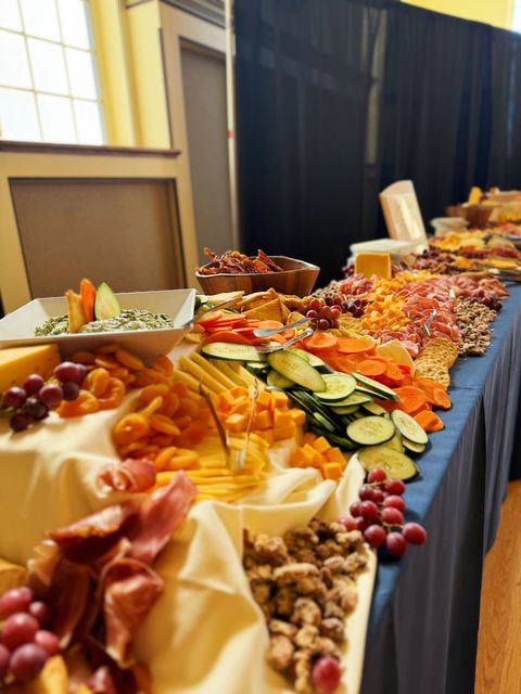 Long buffet table with charcuterie: cheese, grapes, crackers, nuts, vegetables, dips, and meats.