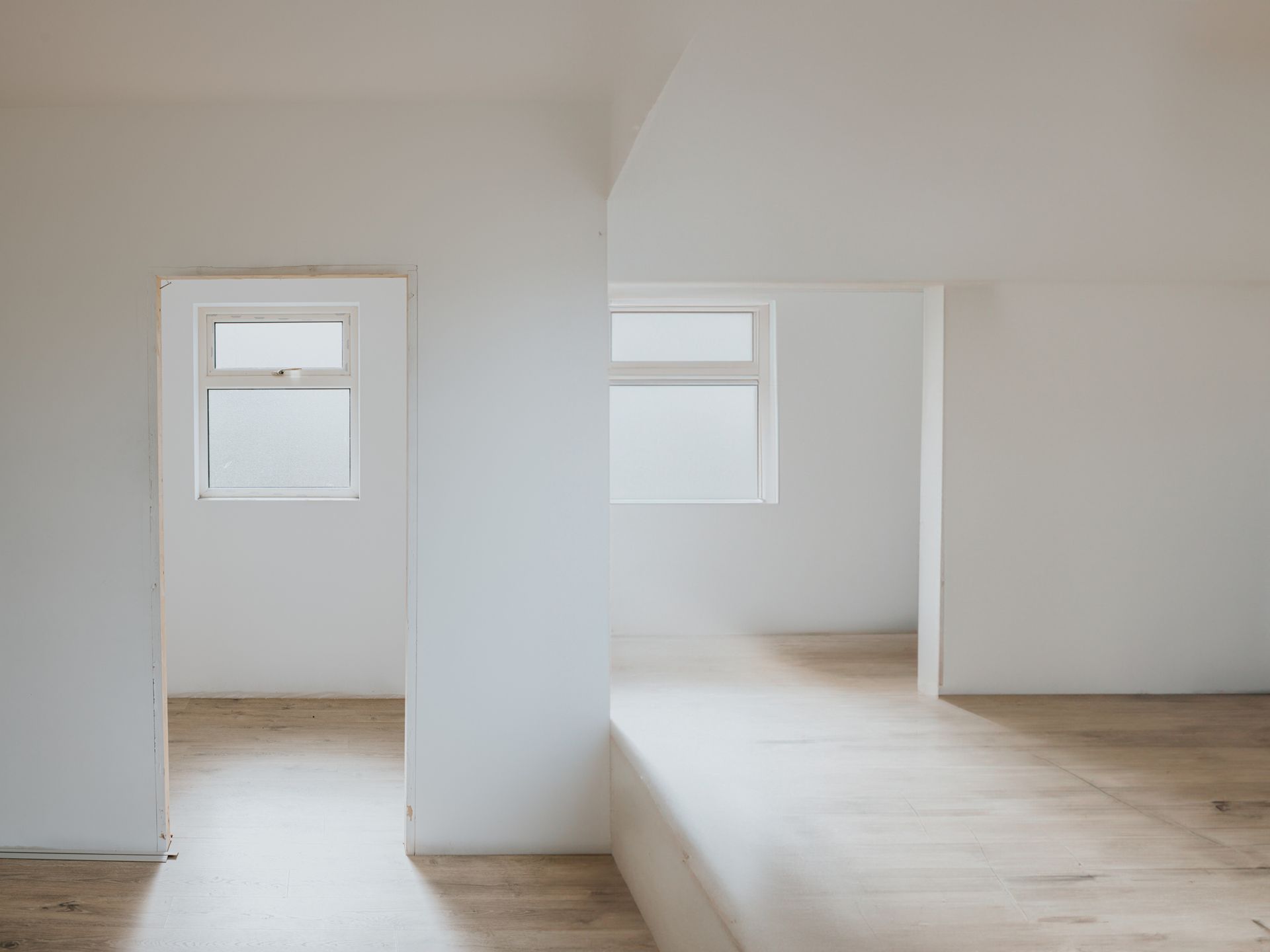 Empty white-walled rooms with two windows and wood-like flooring, bathed in natural light.