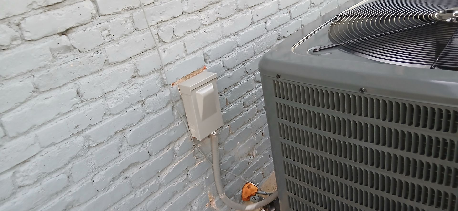 Outdoor HVAC unit beside a white brick wall with a small electrical box and conduit on the wall