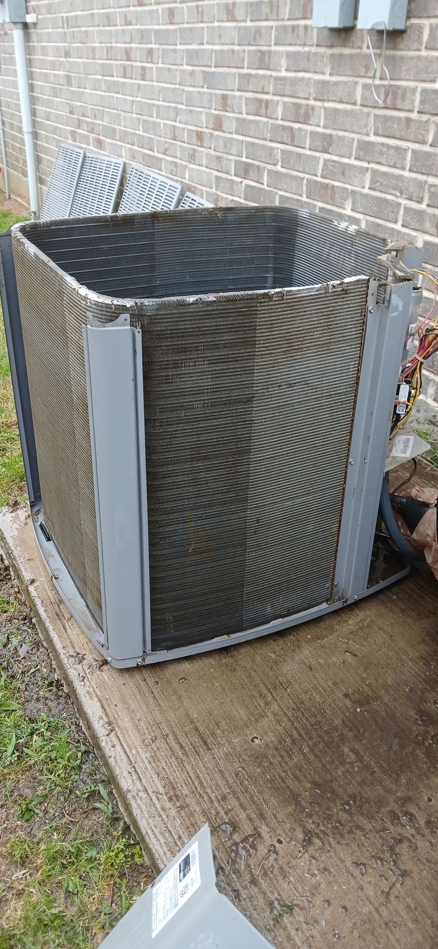 Outdoor HVAC condenser unit beside a brick wall, with metal grille panels and a concrete pad.