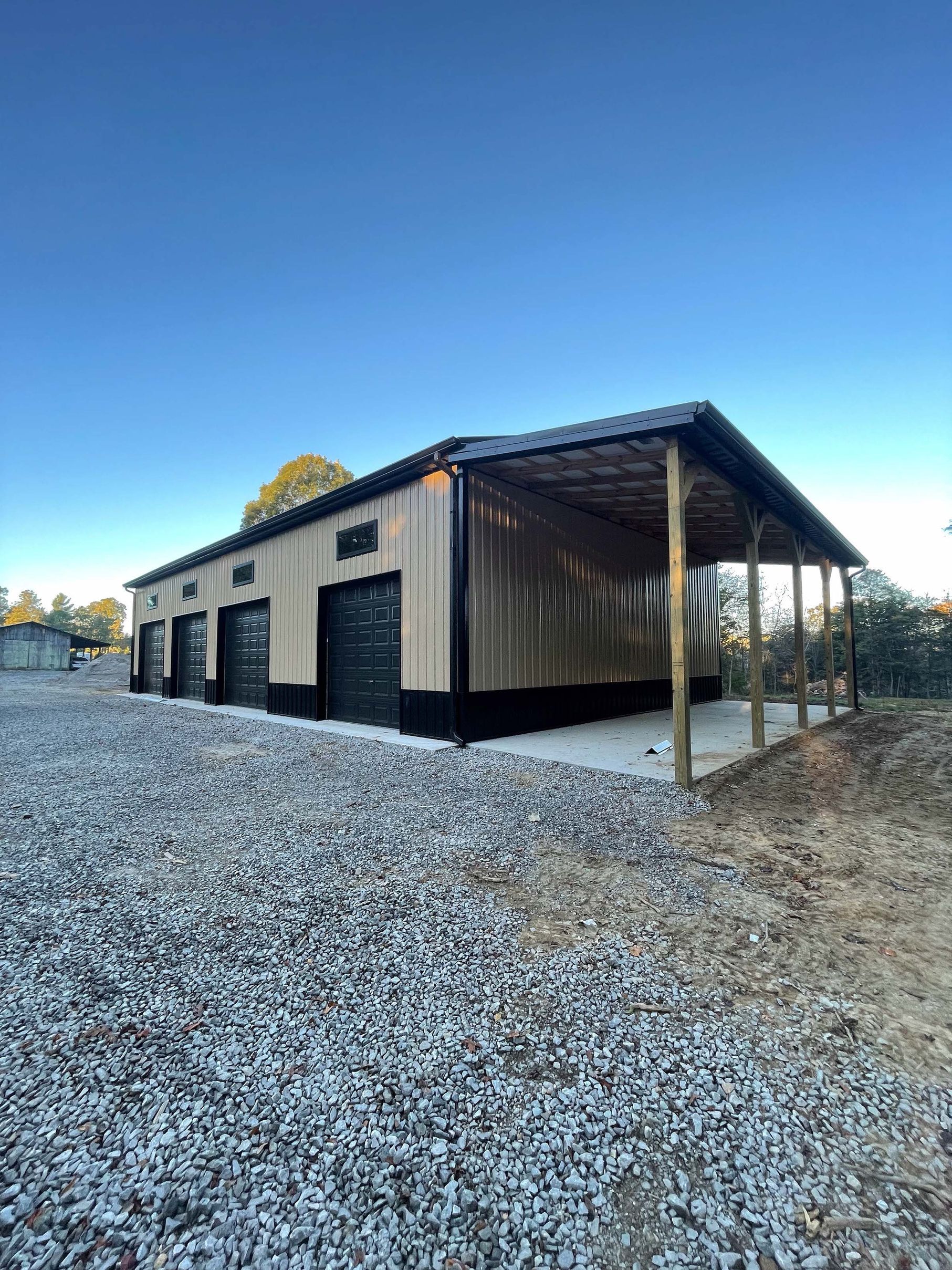 A large metal garage with a covered porch is sitting on top of a gravel lot.