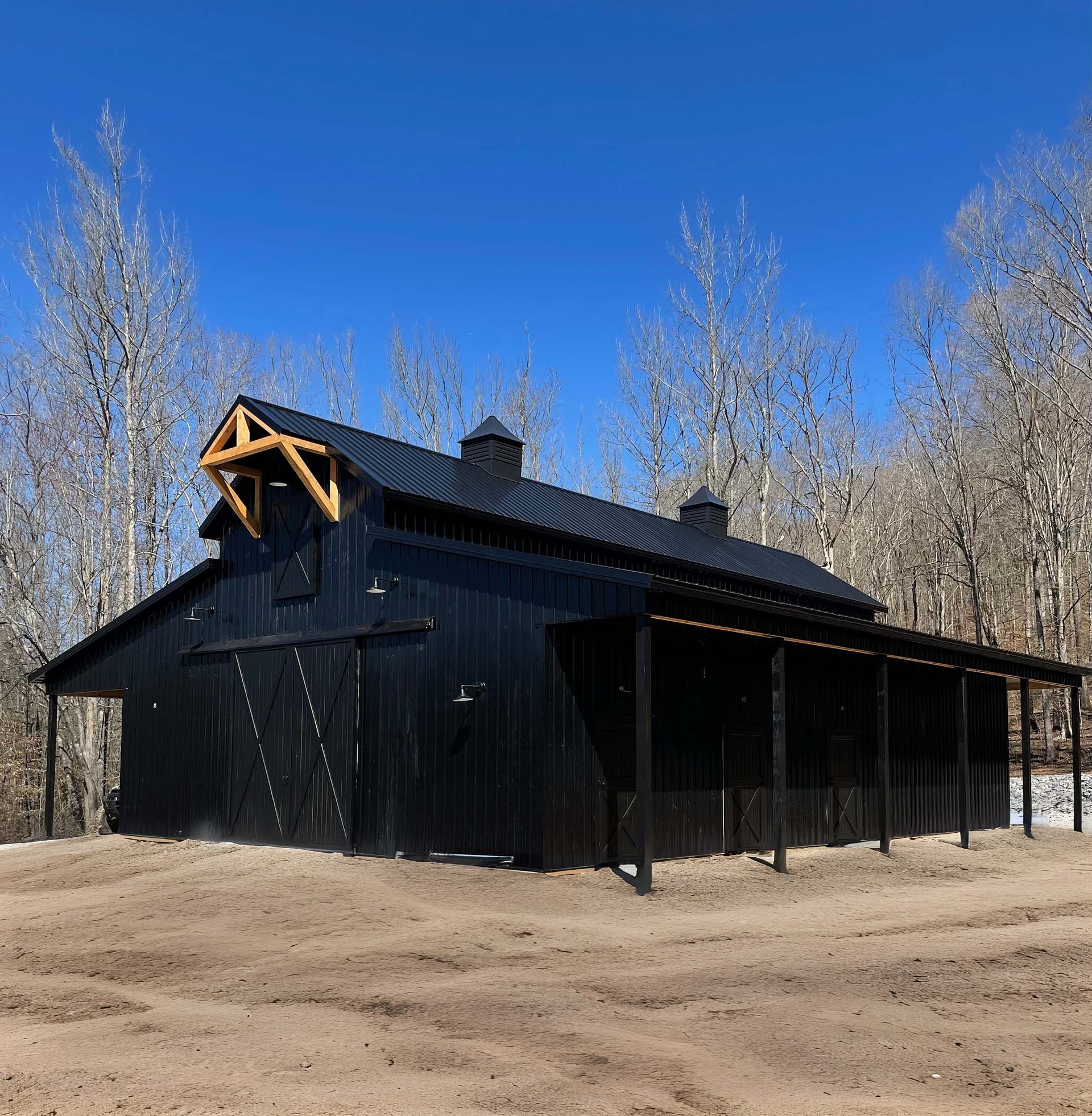 A black barn with a wooden roof is in the middle of a dirt field.