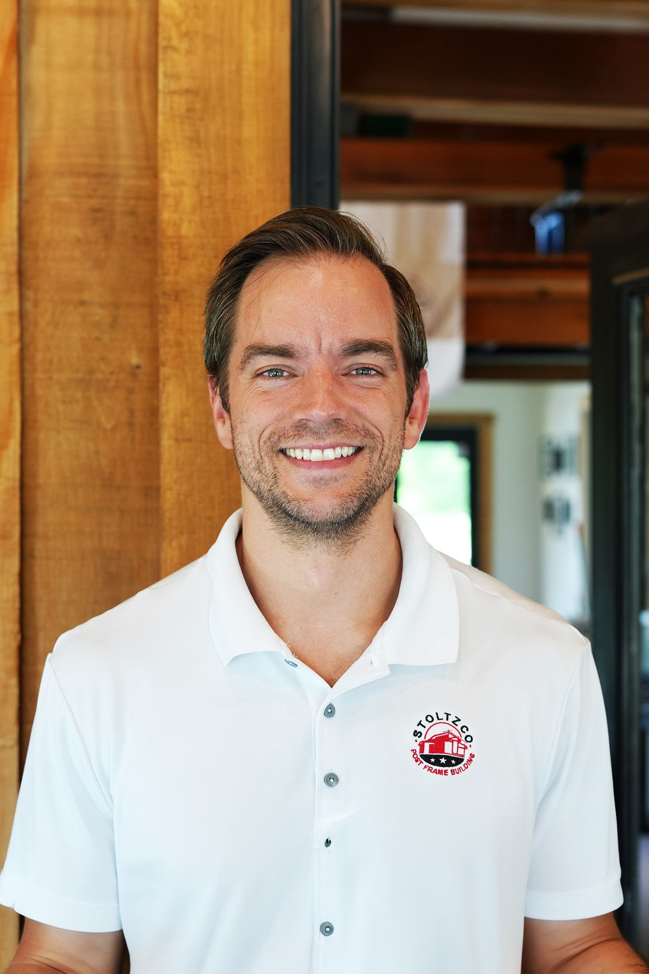A man wearing a black t-shirt is smiling in front of a wooden wall.