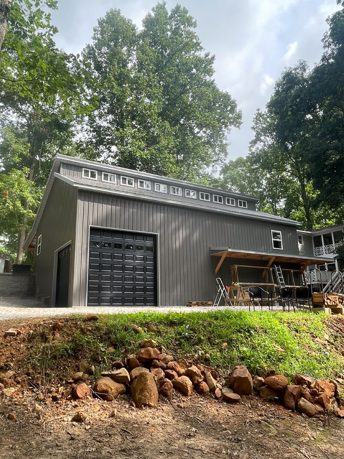 A large gray garage with a black garage door is sitting on top of a hill next to a house.
