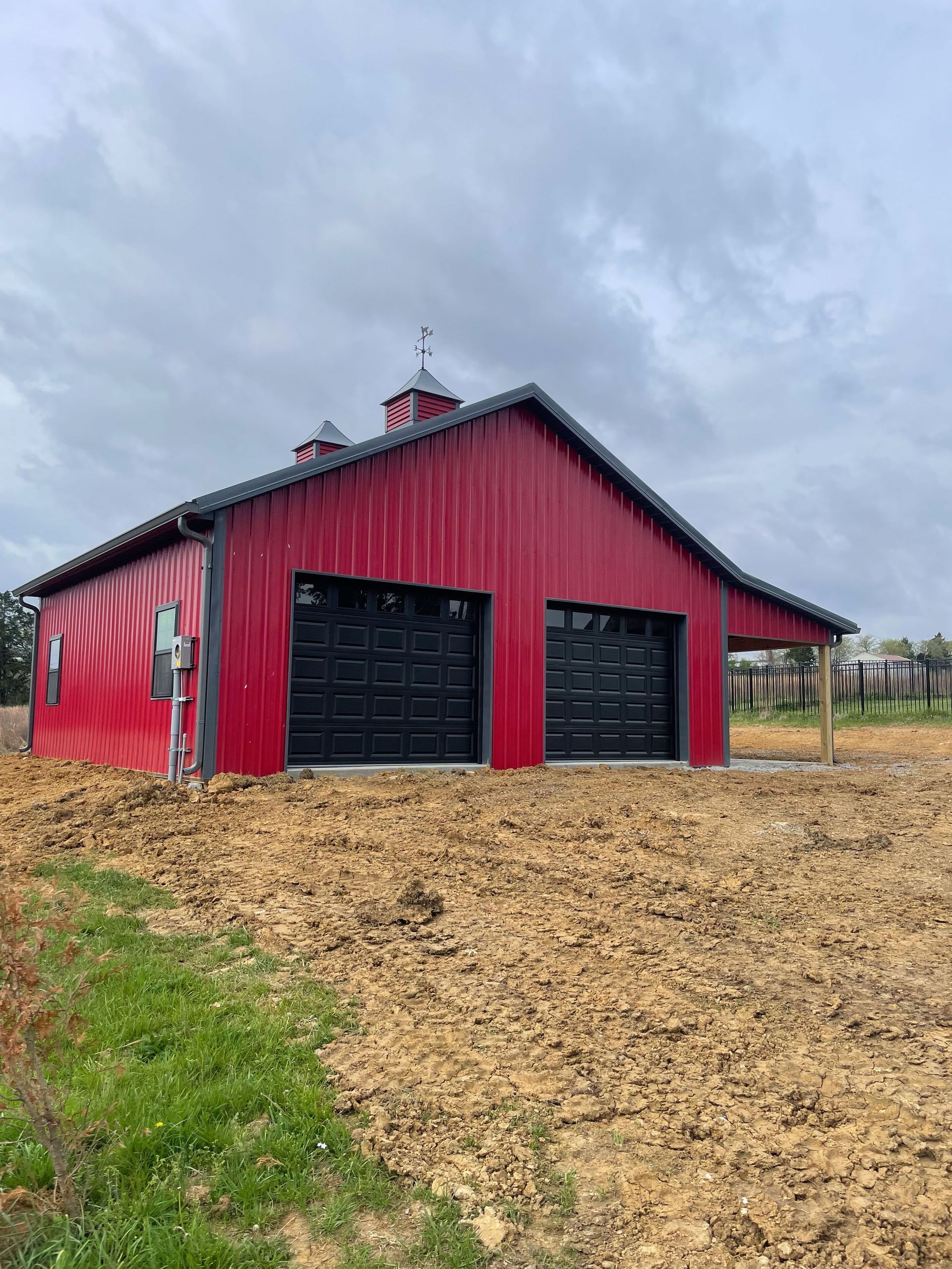 A red barn with black garage doors is sitting in the middle of a dirt field.