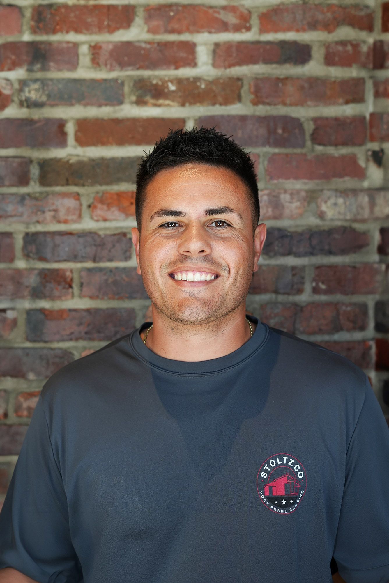 A man in a black polo shirt is smiling in front of a wooden wall