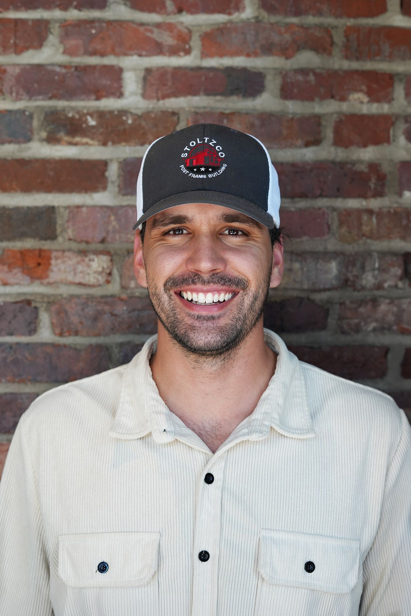 A man wearing a hat and a brown shirt is smiling in front of a wooden wall