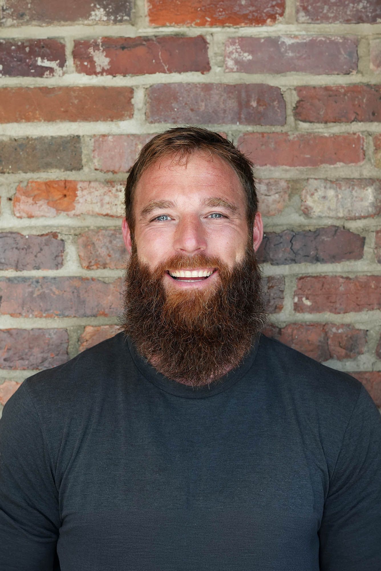 A man with a long beard is smiling in front of a wooden wall.