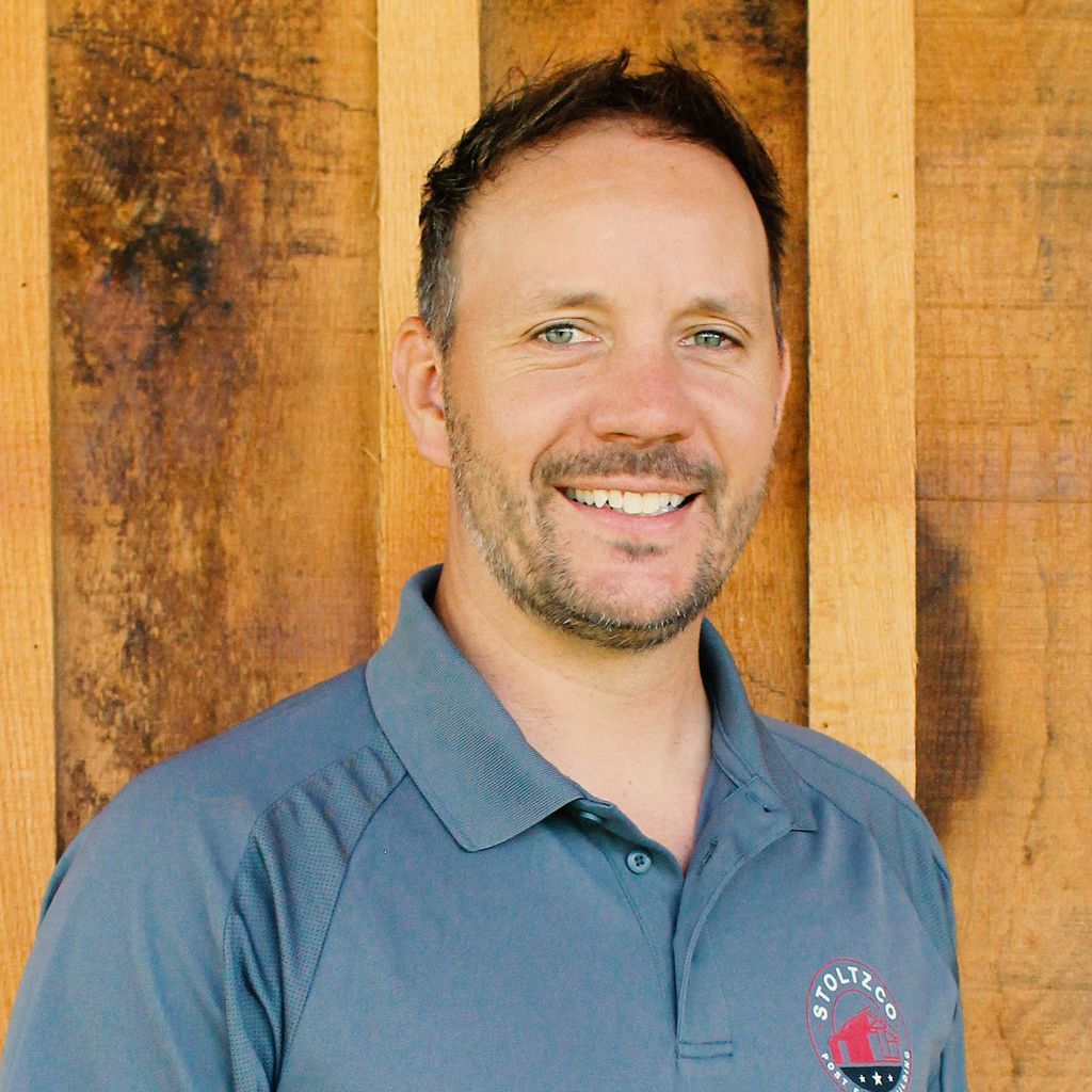 A man with a beard is smiling in front of a wooden wall.