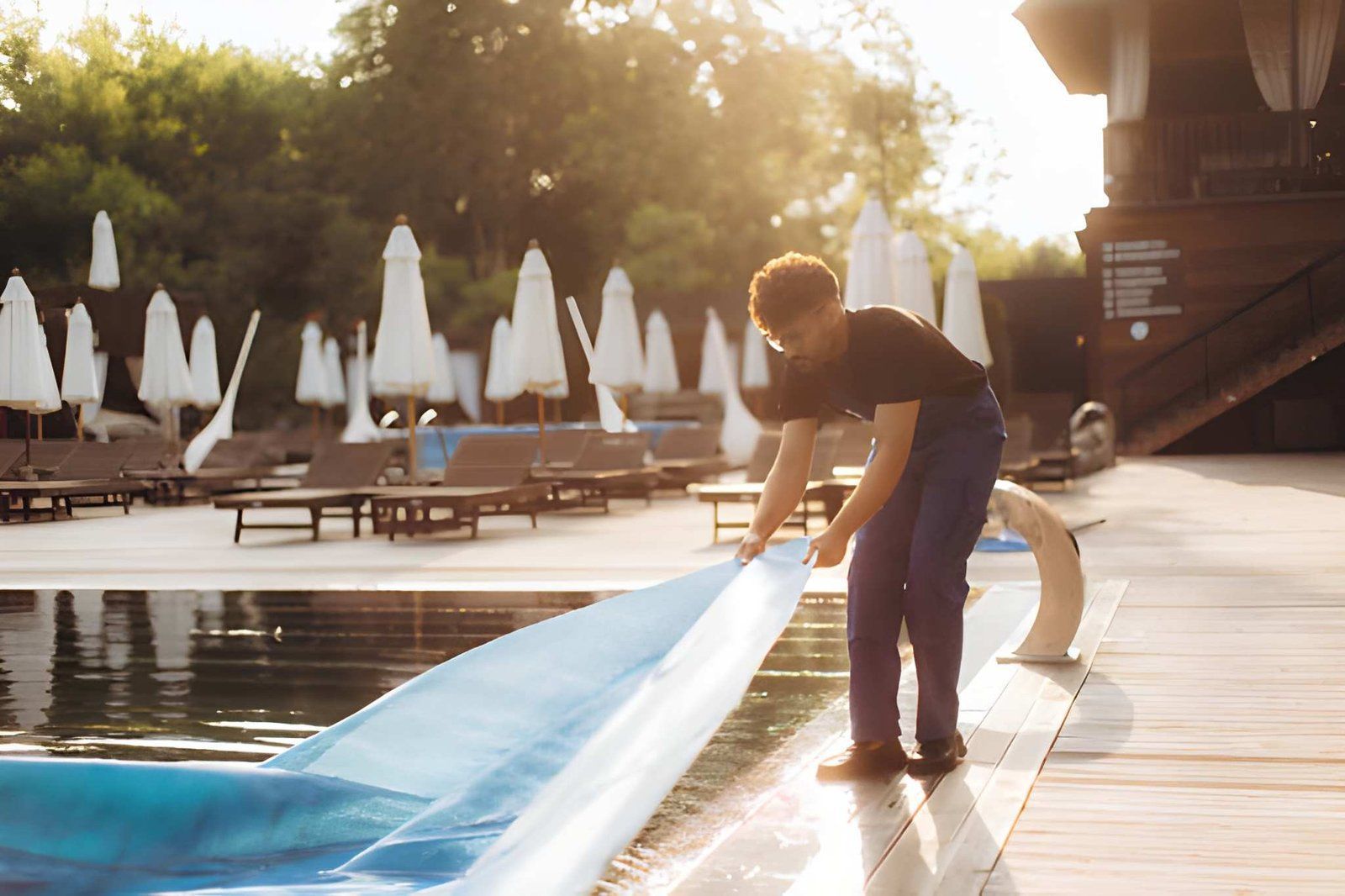 Person unrolls a blue pool cover beside a sunlit outdoor swimming pool with lounge chairs and umbrellas.