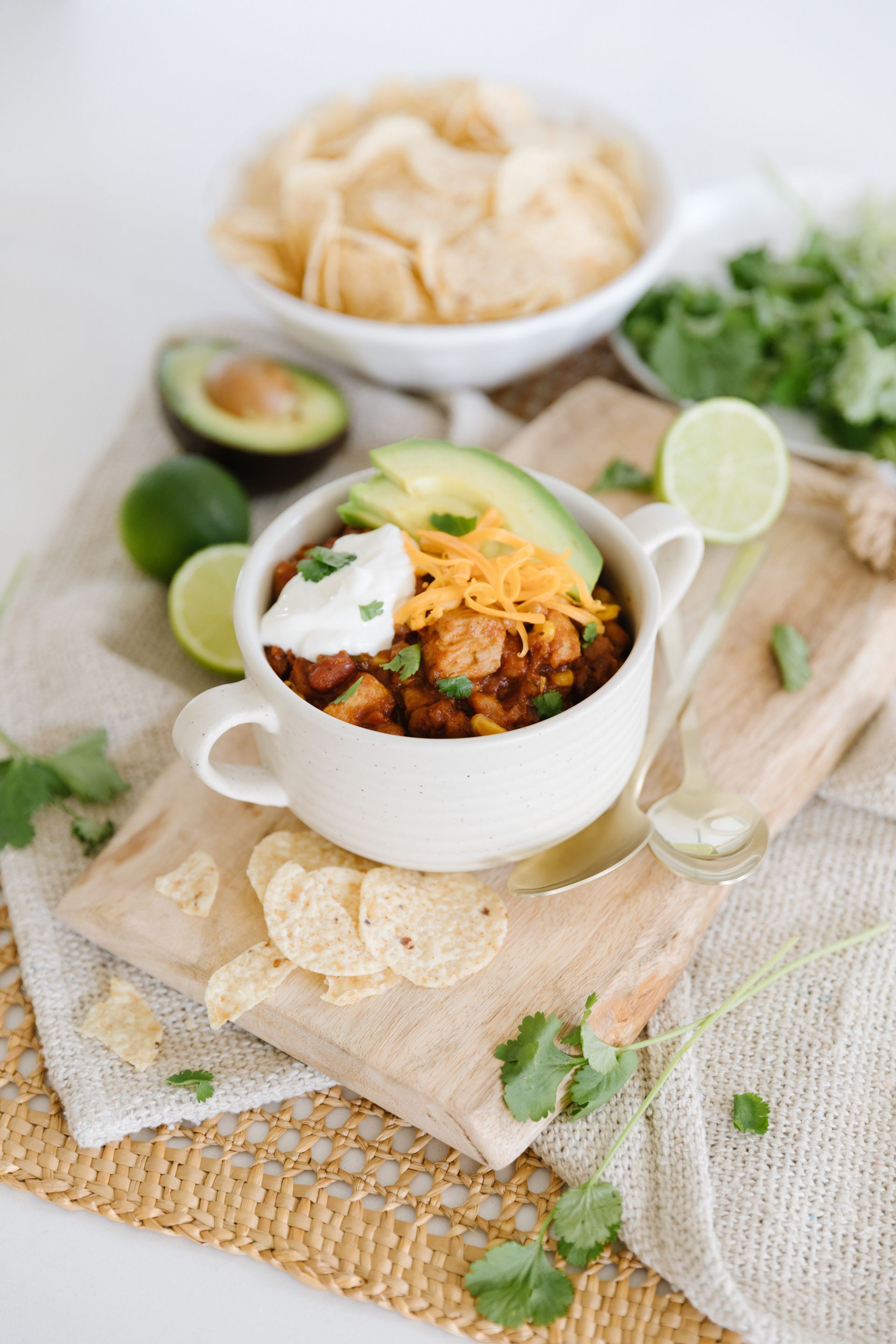 a bowl of chili with sour cream , cheese , avocado and chips on a wooden cutting board .