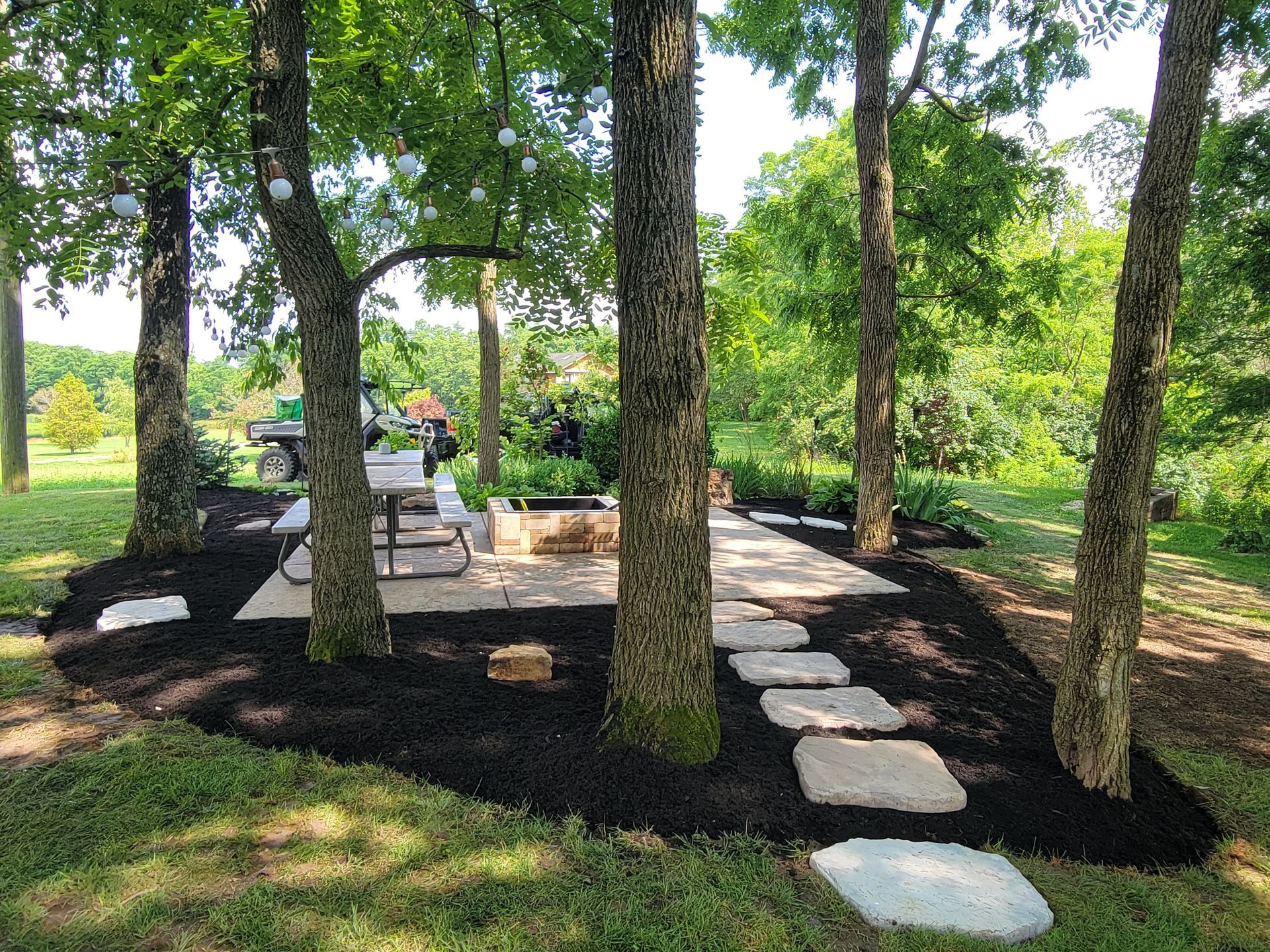 A stone pathway leads to a patio with a fire pit, surrounded by trees and mulch.