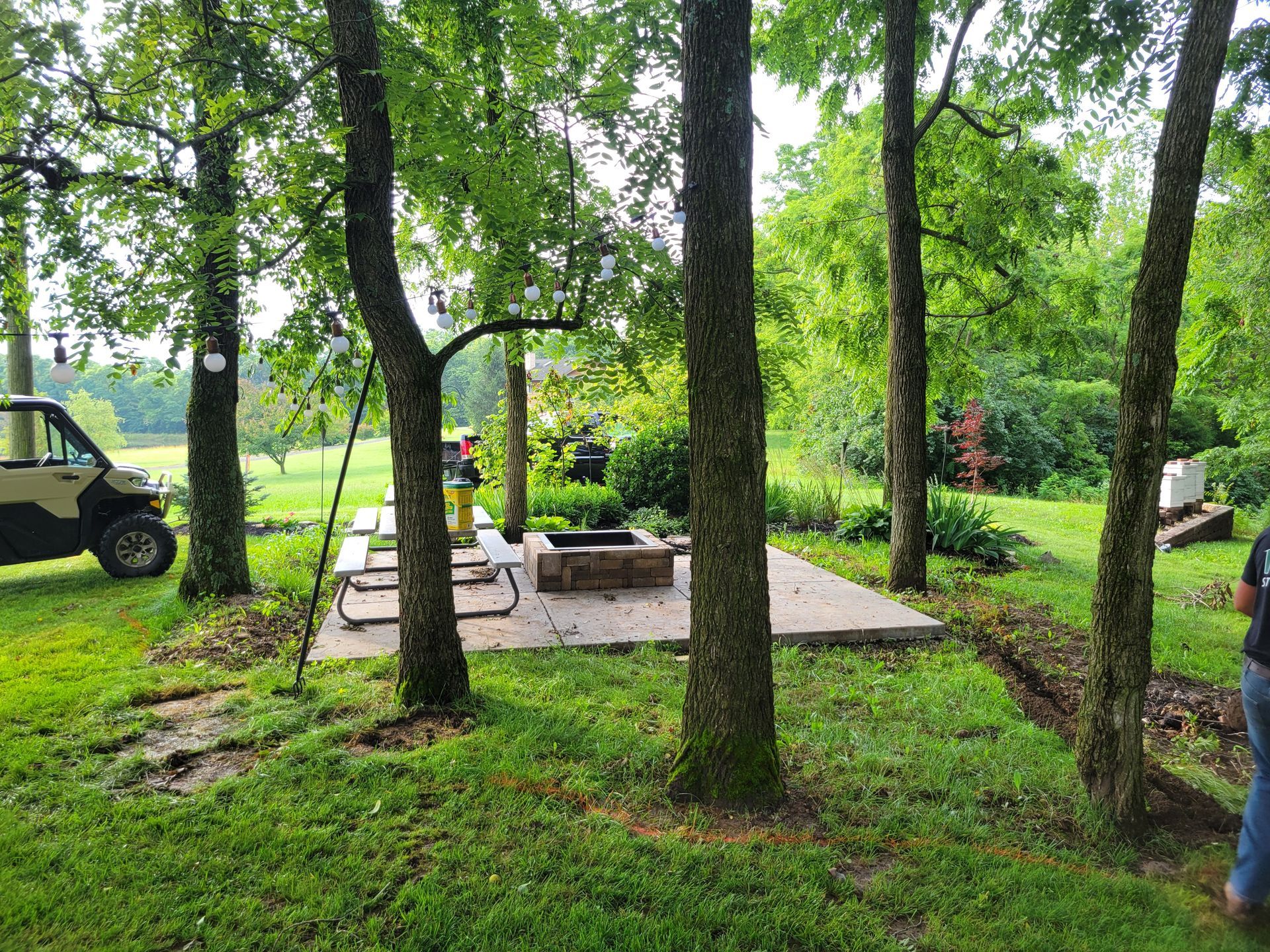 Picnic area with fire pit and table, framed by trees, grassy lawn. Utility vehicle on the left.