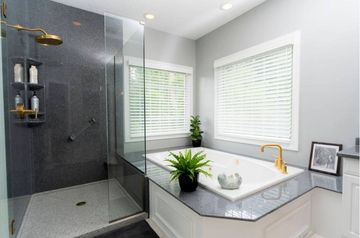Modern bathroom with black glass shower, white tub, gray counters, and gold faucet beneath window blinds