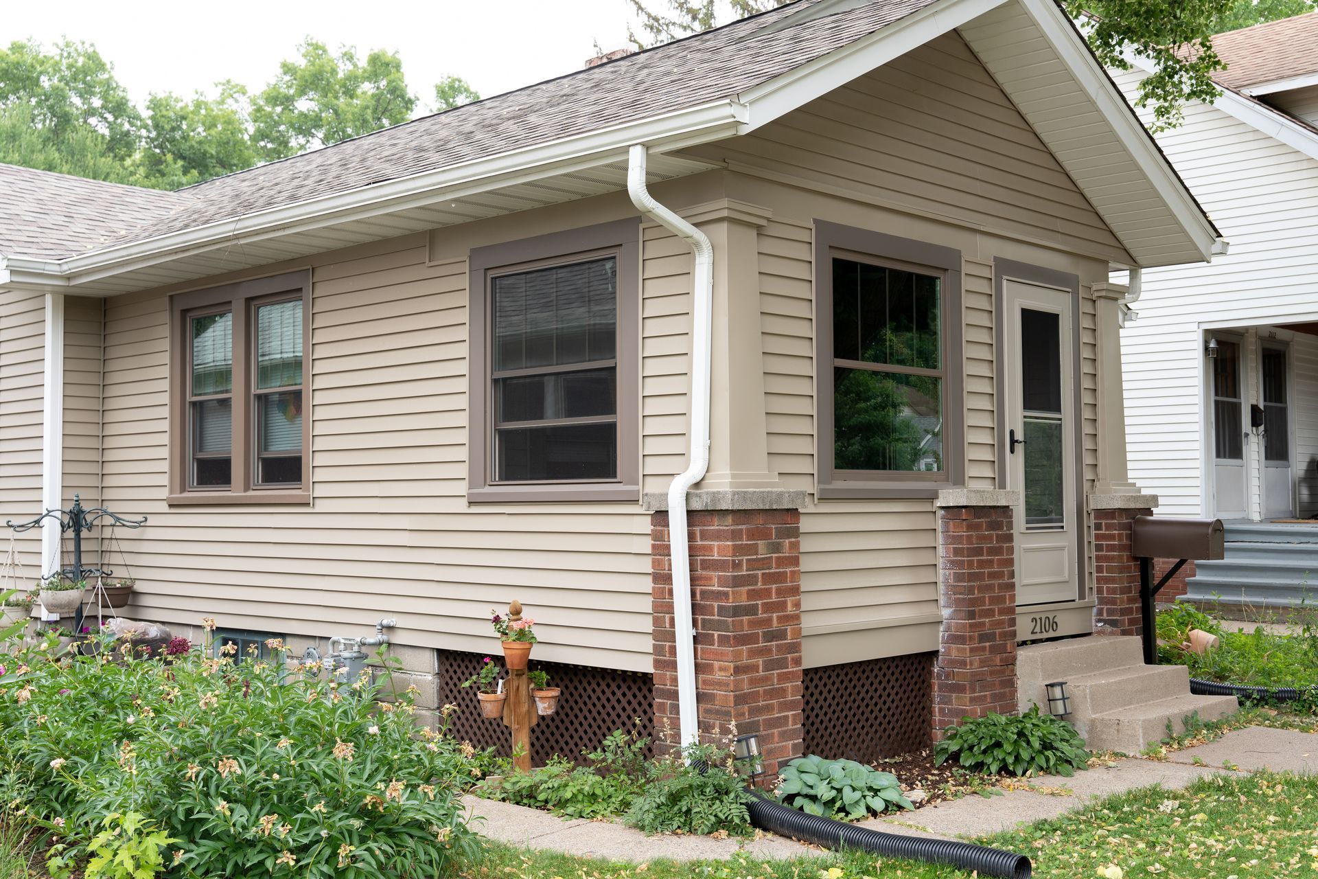 Small beige house with brick foundation, windows, and a front stoop surrounded by greenery