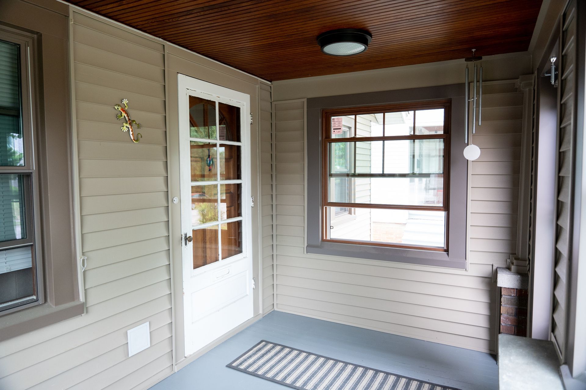 Covered porch with beige siding, white door, brown-trimmed window, and wood-paneled ceiling.