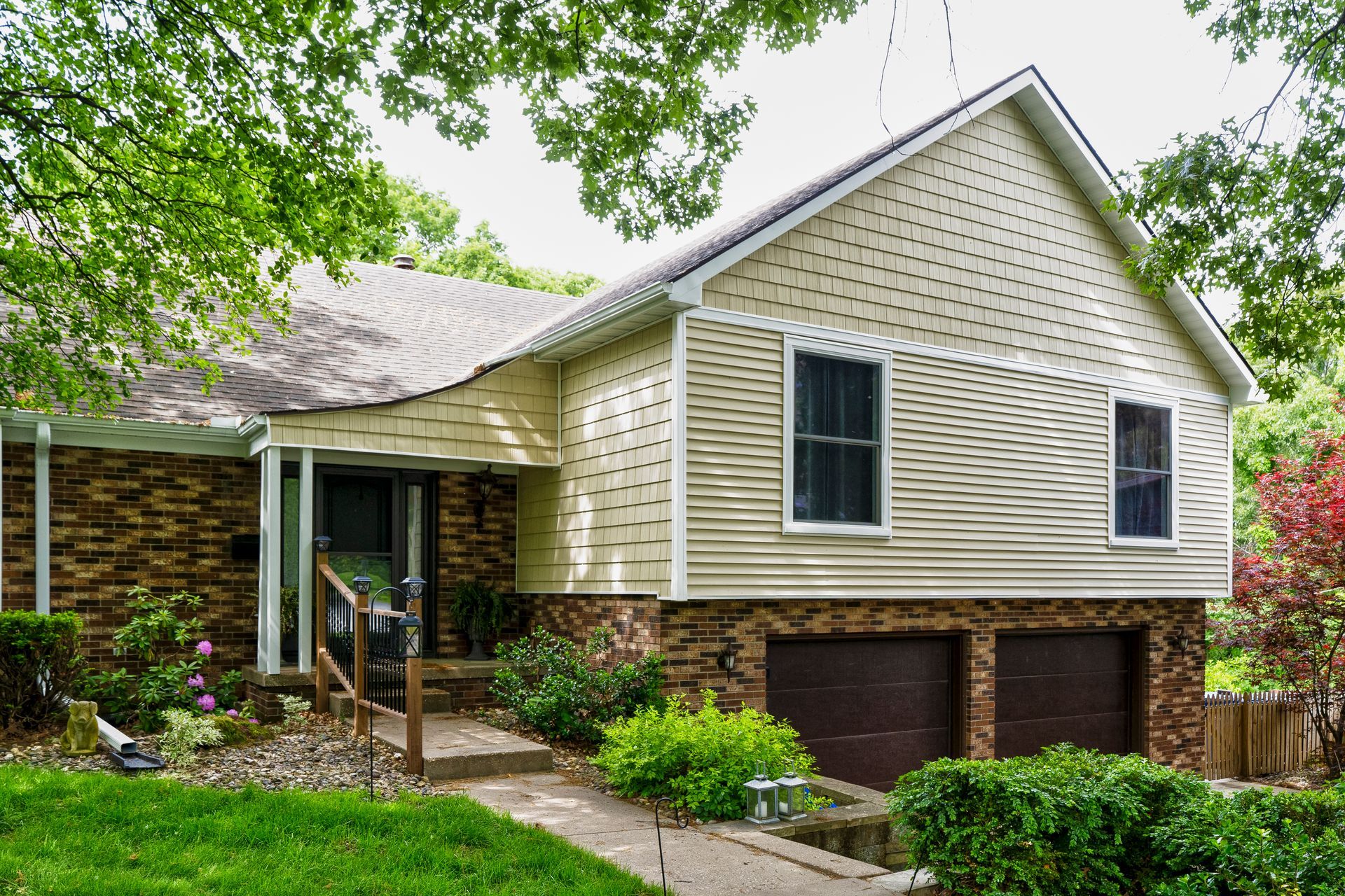 Suburban house exterior with beige siding, brick base, and a front walkway surrounded by greenery