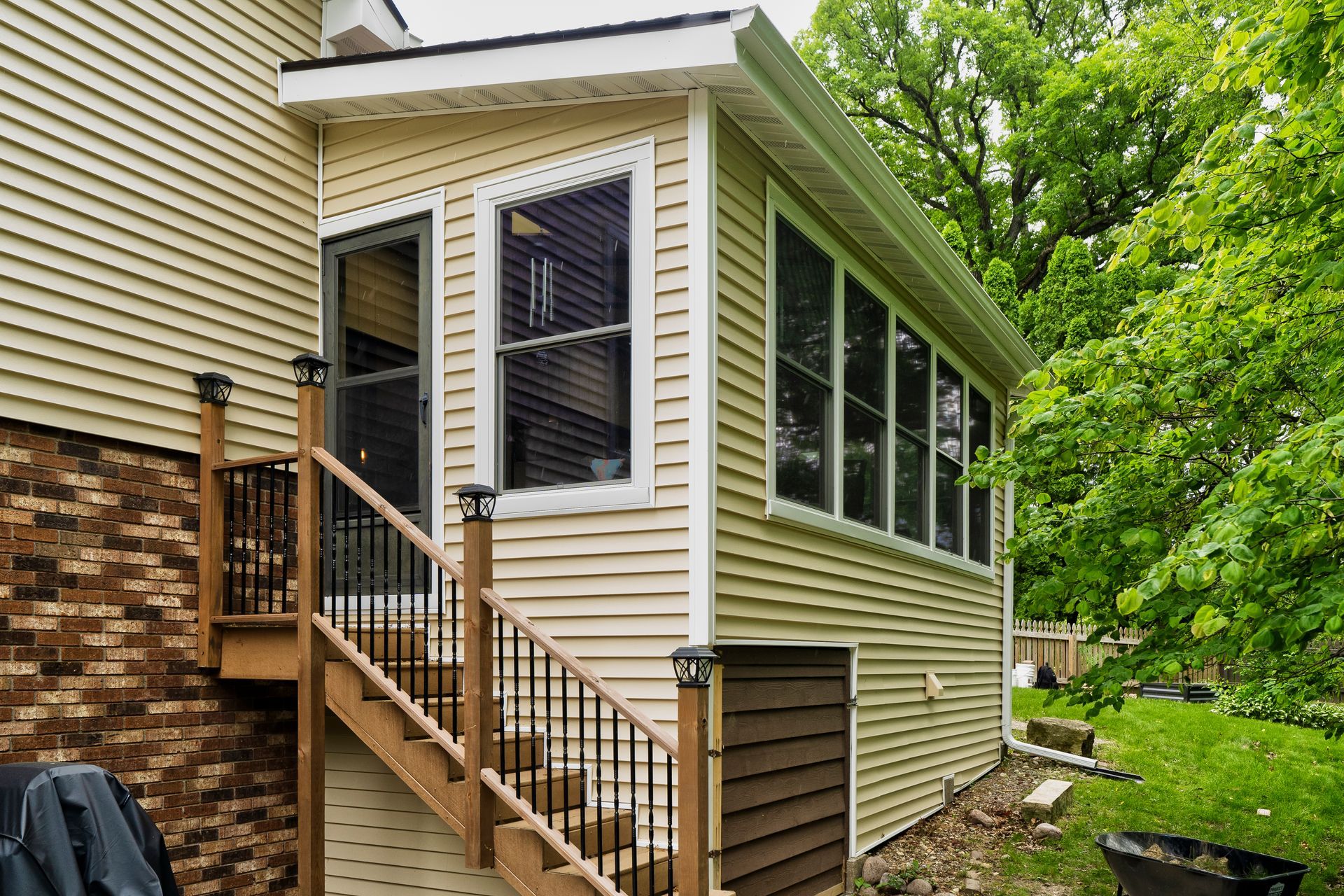 Enclosed sunroom addition with beige siding, windows, and exterior stairs beside a house