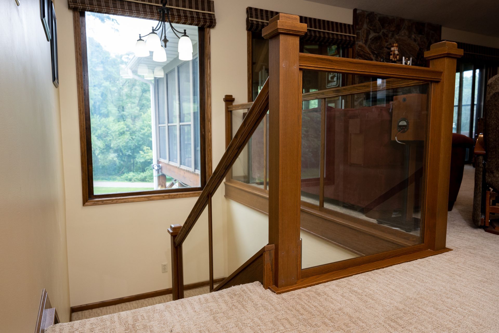 Wooden indoor stair lift with glass safety panel beside a carpeted staircase and window.