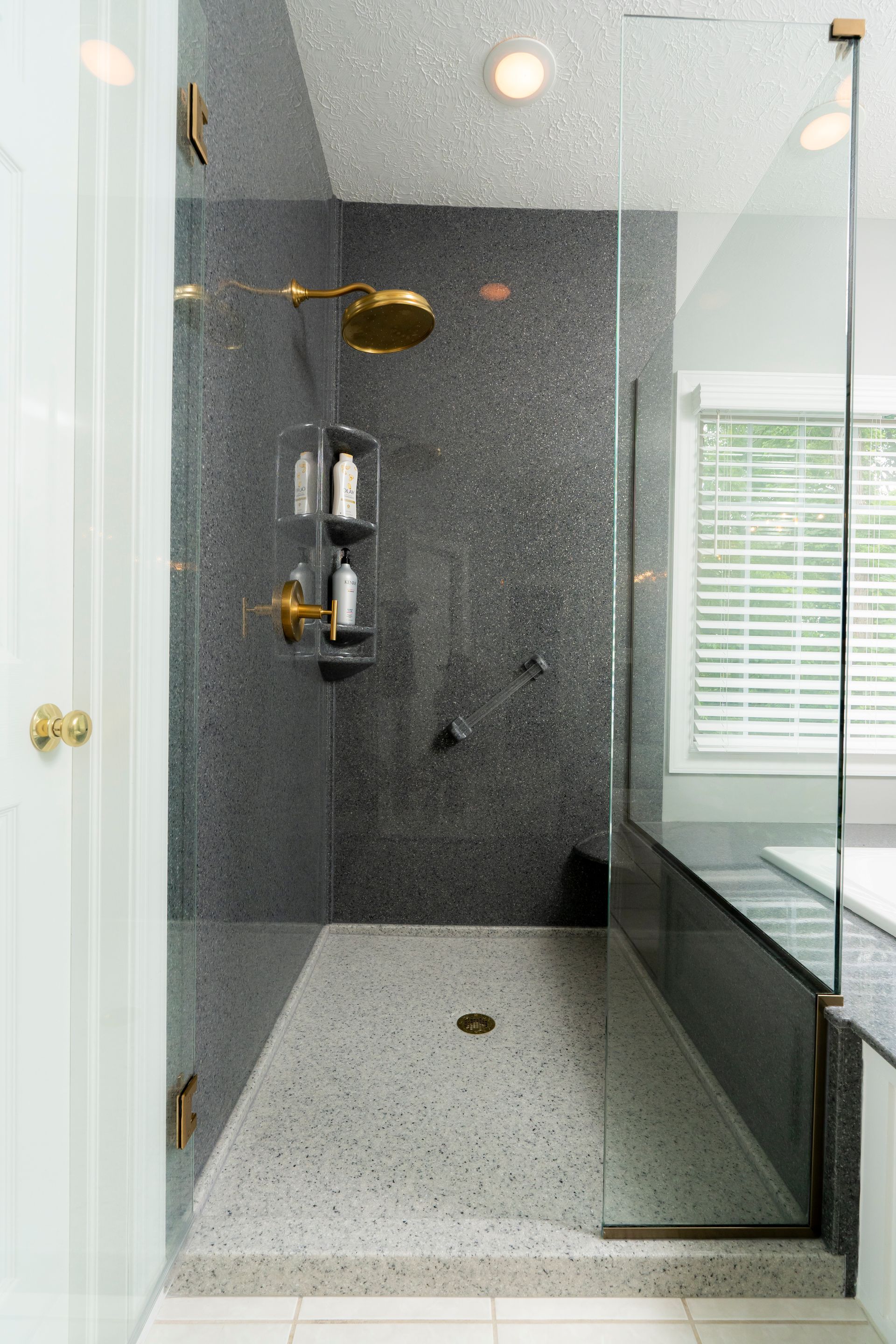 Modern walk-in shower with glass enclosure, gray tile walls, brass fixtures, and pebble-textured floor.