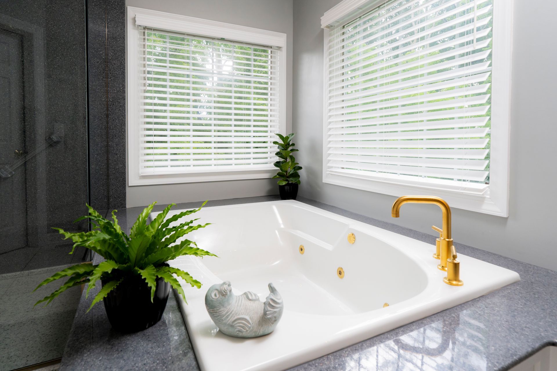 Bright bathroom with a white soaking tub, gold faucet, gray tile, and potted plants by the windows.