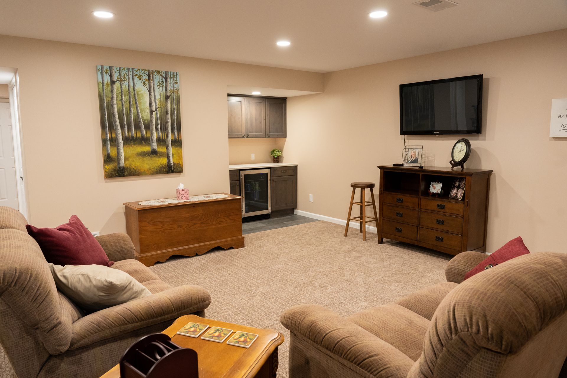 Cozy basement living room with beige sofas, wooden TV stand, small window, and forest wall art