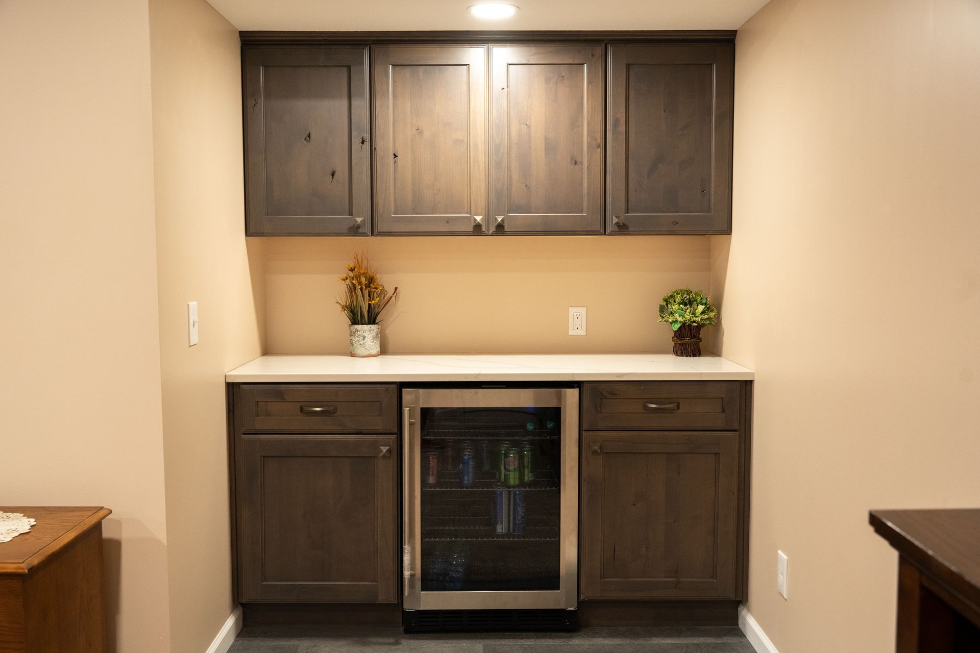 Built-in dark wood bar cabinet with mini fridge, white countertop, and potted plants on either side.
