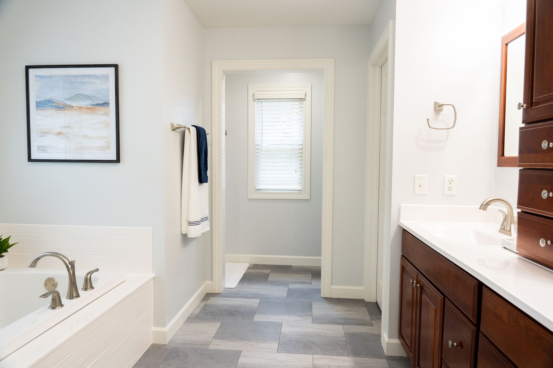 Bright bathroom with a white tub, dark wood vanity, blue-gray tile floor, and window at the end of the hall