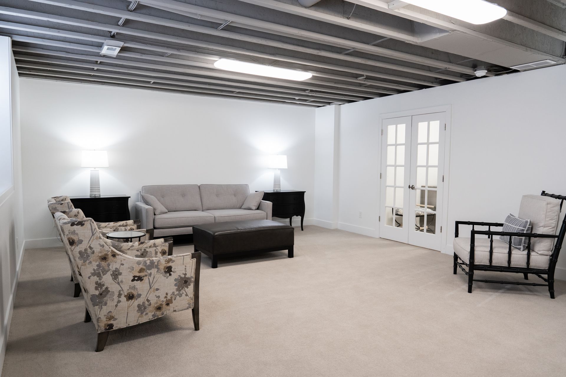 Spacious living room with beige carpet, gray sofa, floral chair, black table, and white walls and ceiling beams
