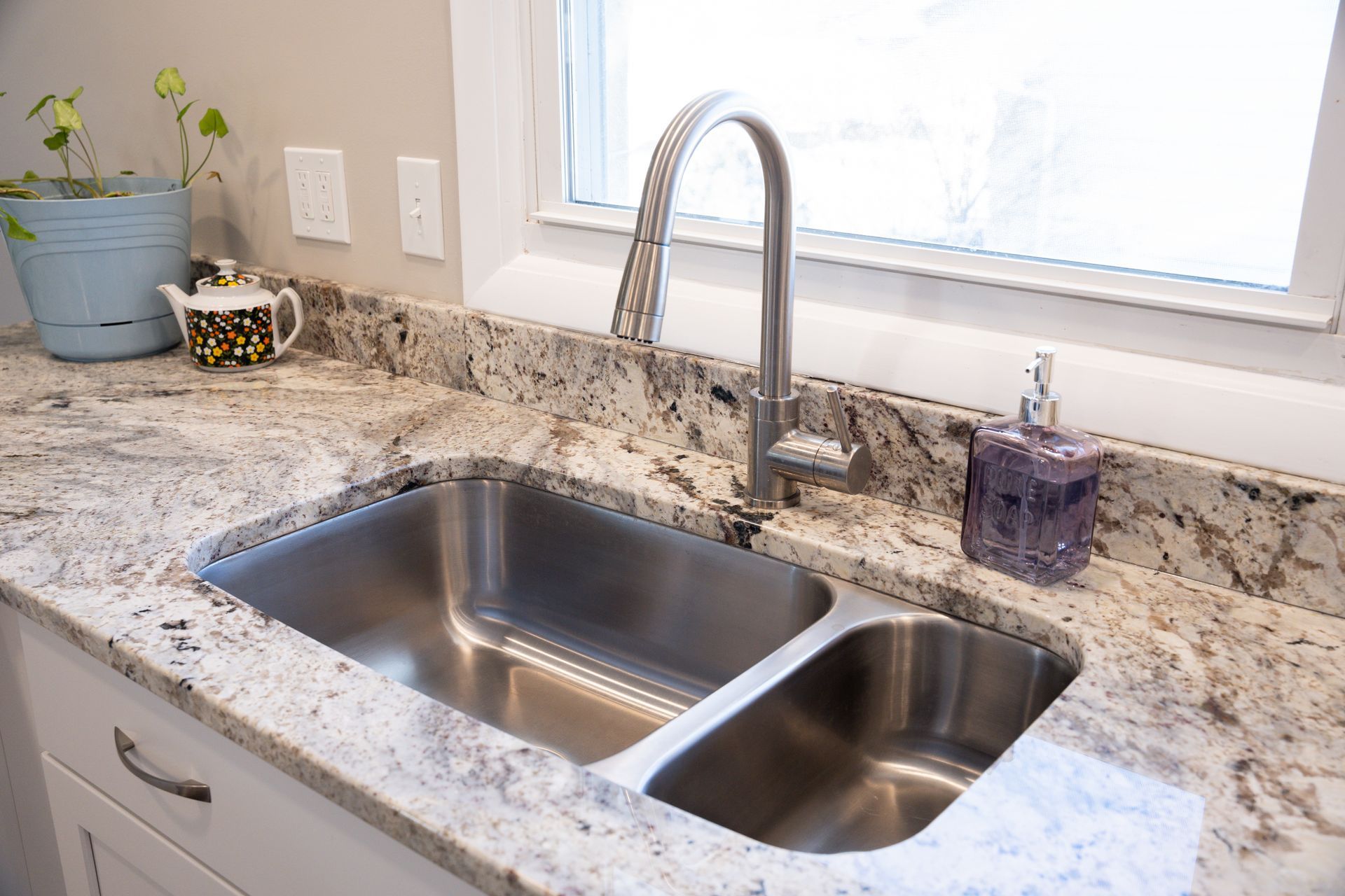 Granite kitchen countertop with double stainless-steel sink, faucet, and small potted plants by a window